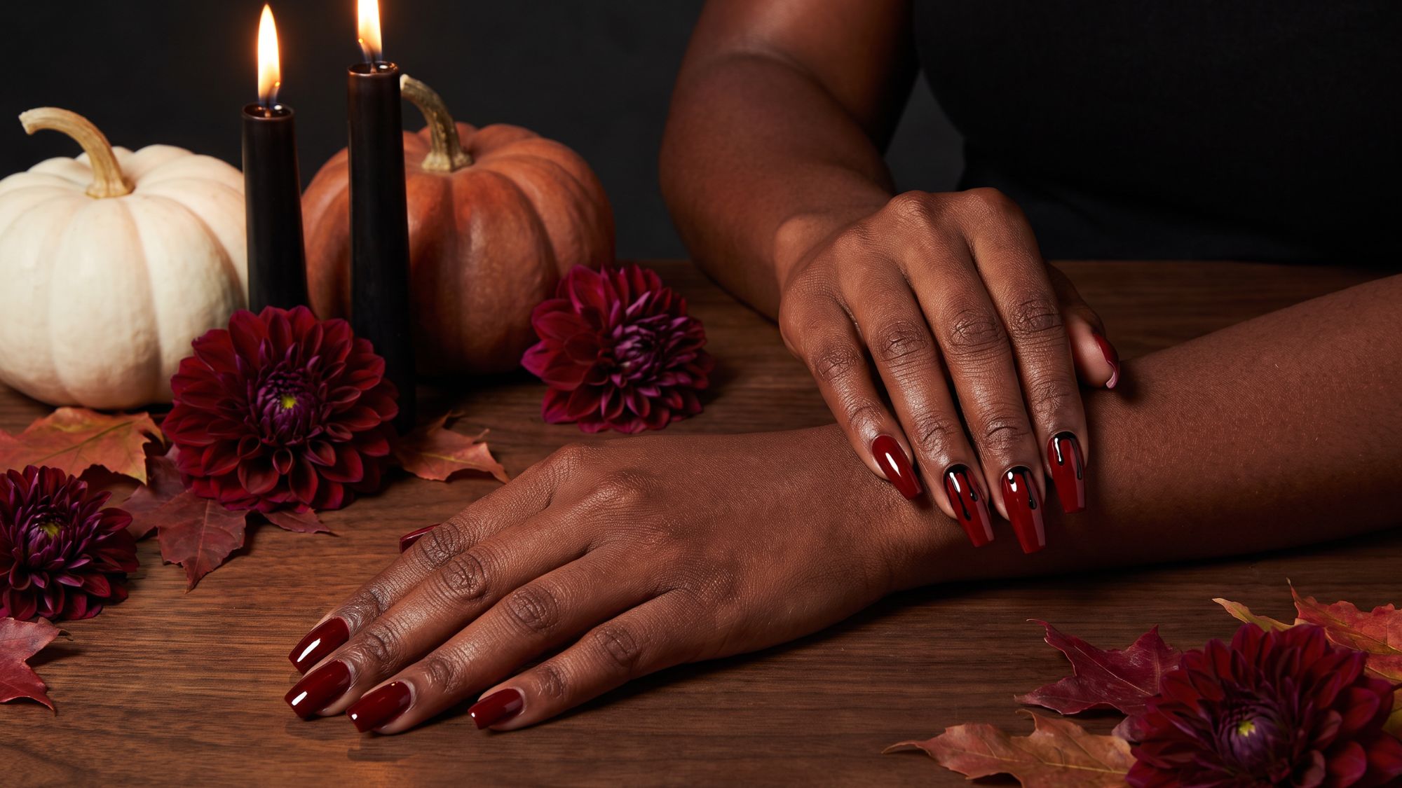 Two hands with Halloween manicures, short oxblood square nails and long blood red coffin nails with black drip detail, staged with black candles and heirloom pumpkins
