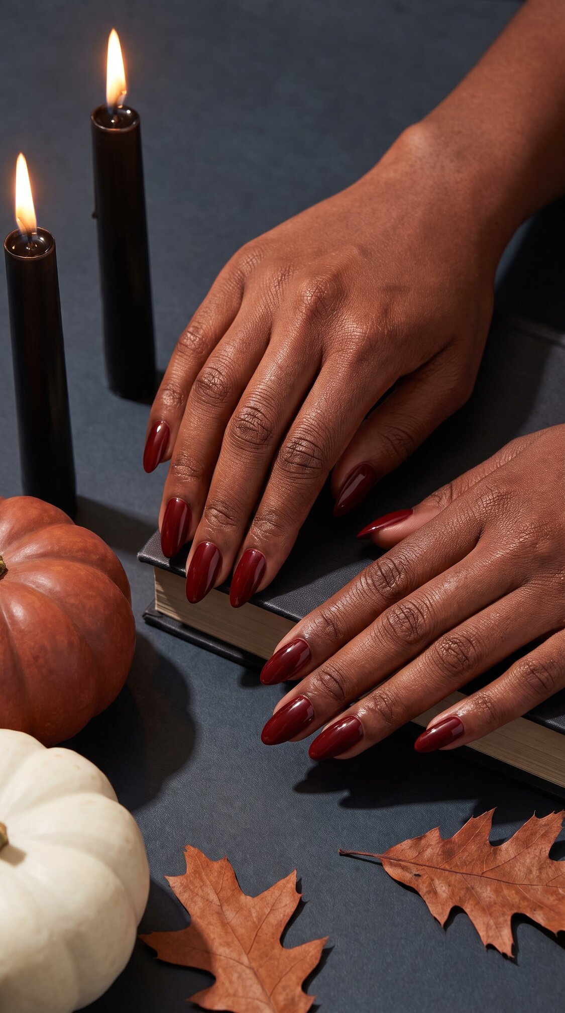 A person with long, dark red halloween nails acrylic rests their hands on a book beside black candles, a white and orange pumpkin, and autumn leaves on a dark surface.