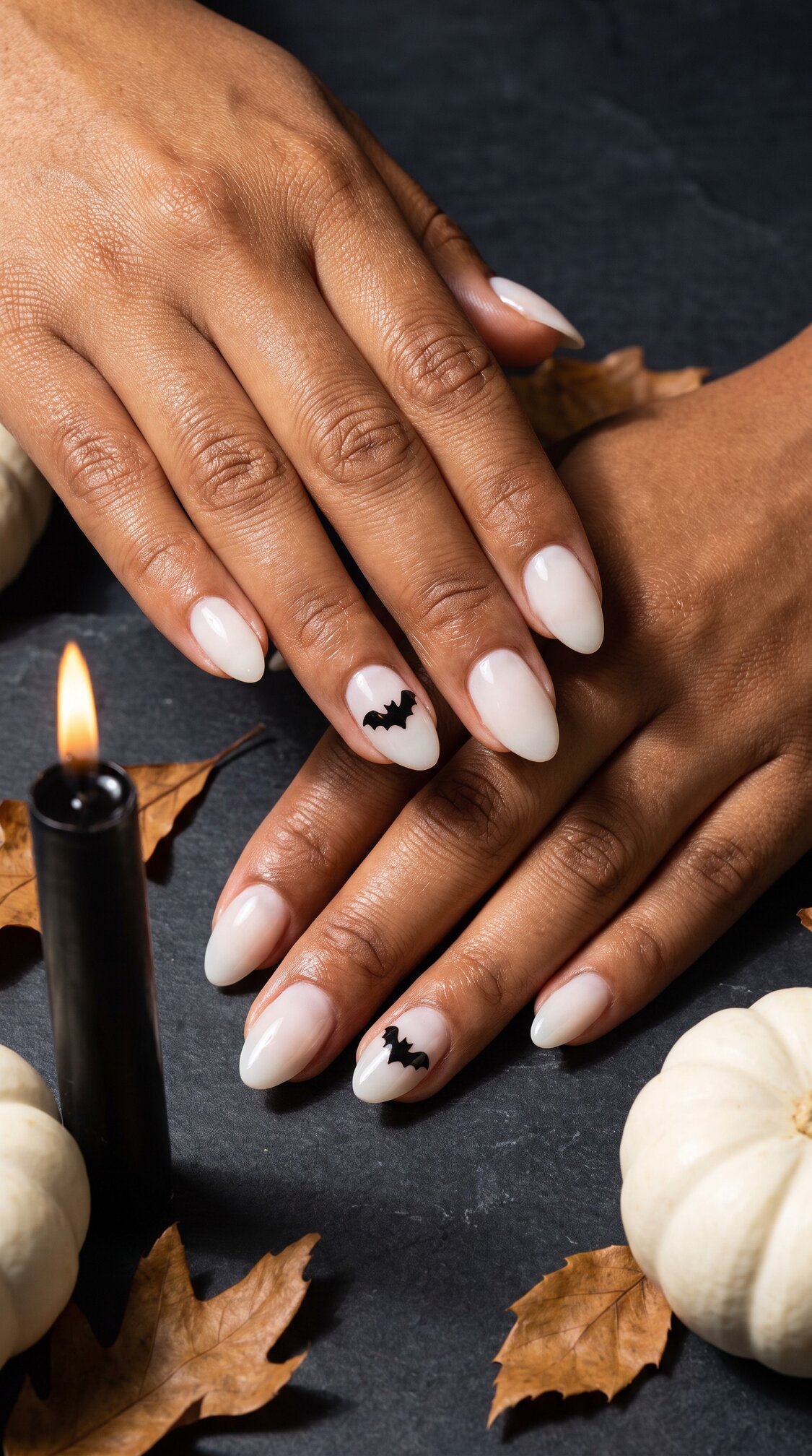 Hands with almond-shaped, pale halloween nails acrylic, some featuring small black bat designs. The hands rest on a dark surface with white pumpkins, brown leaves, and a lit black candle nearby.