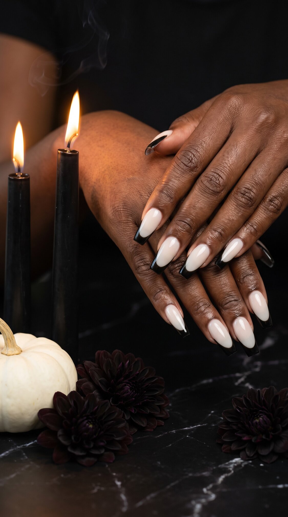 A person with nude and black-tipped halloween nails acrylic manicure rests their hands on a dark marble surface near two lit black candles, dark flowers, and a small white pumpkin.