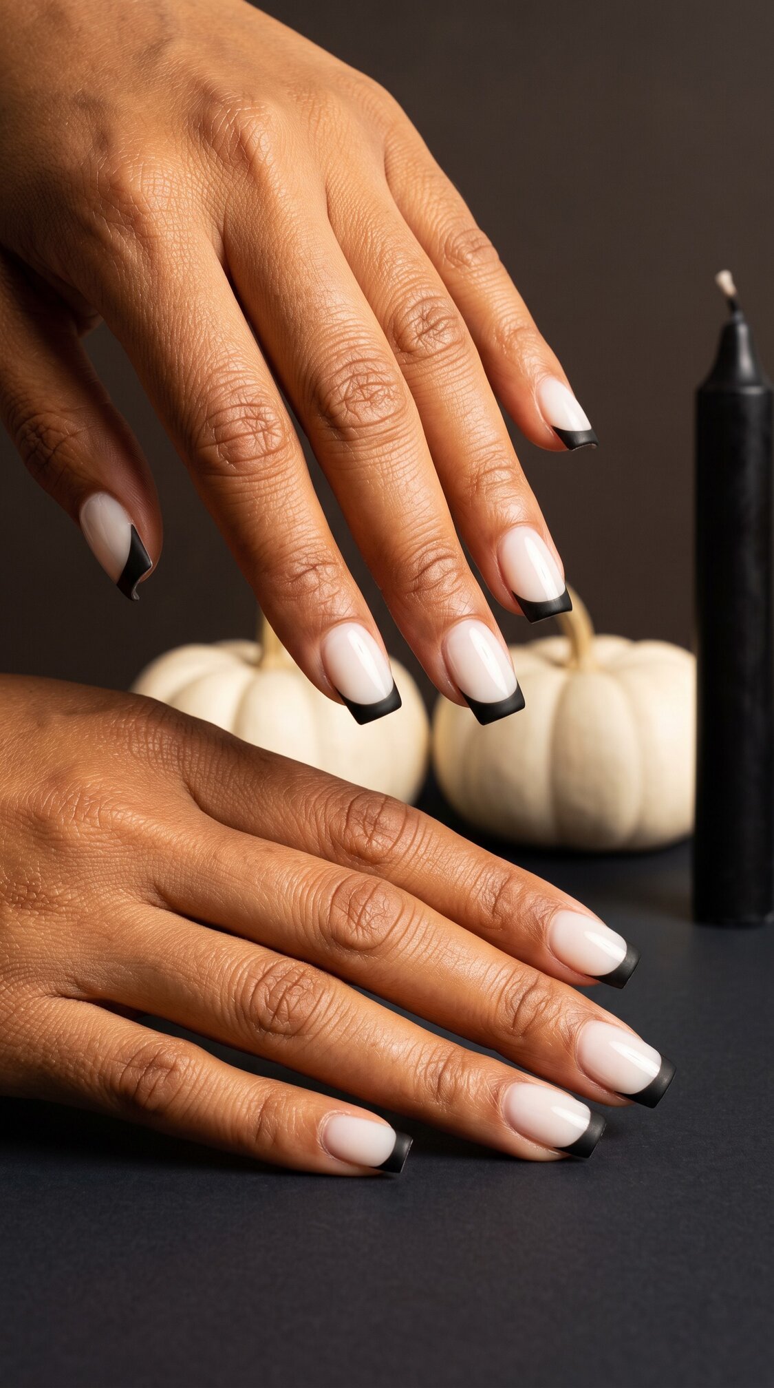 Two hands with almond-shaped halloween nails acrylic, featuring a white base and black tips. A white pumpkin and a black candle are in the background, set against a dark surface.