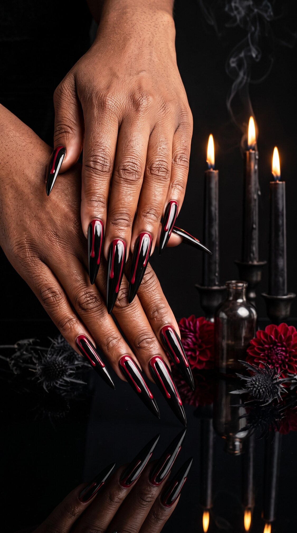 Hands with long, pointed black and red halloween nails acrylic are displayed in front of lit black candles, red flowers, and dark vases, creating a dramatic and gothic atmosphere.
