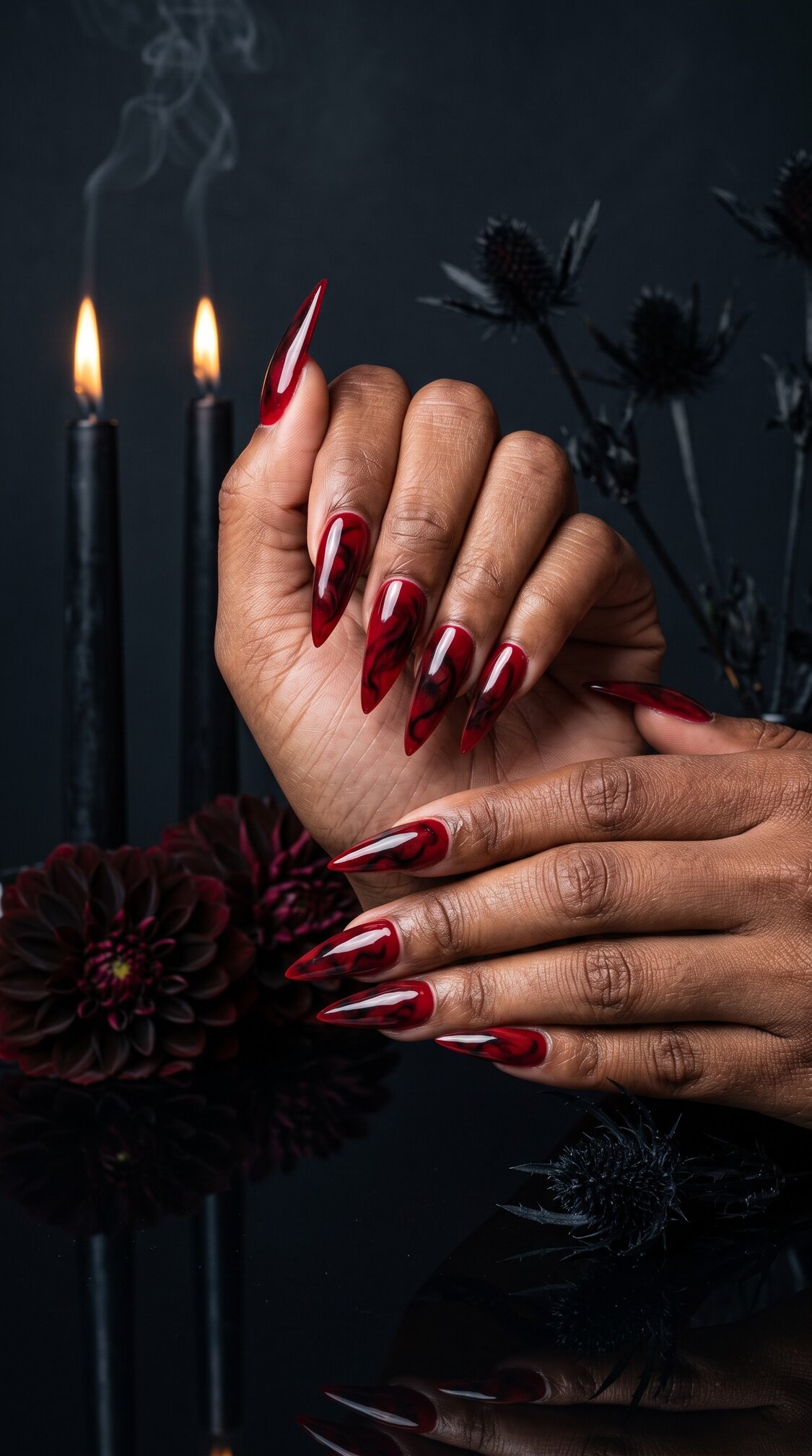A person with long, pointed red and black ombre halloween nails acrylic poses hands together. Black candles, dark flowers, and a black background add a gothic, dramatic mood.