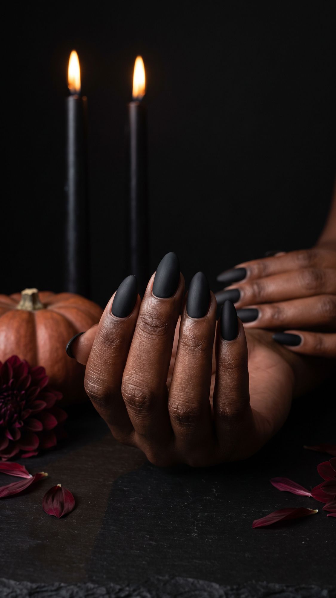 A hand with long matte black nails—perfect for those seeking Glam Halloween Nails—is displayed near a small pumpkin, dark flowers, and two lit black candles, all set against a black background.