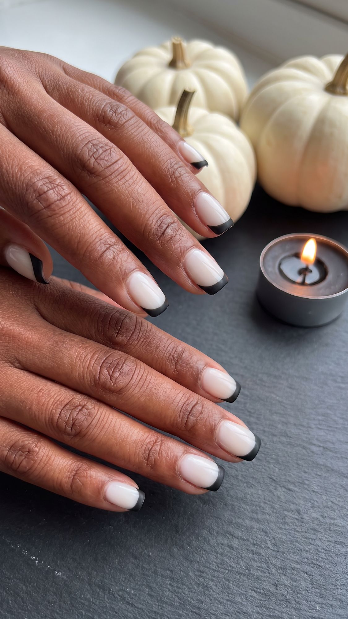 Hands with a black French manicure rest on a dark surface near a lit candle and two small white pumpkins, creating an elegant and autumn-inspired scene perfect for showcasing chic Halloween Nails.