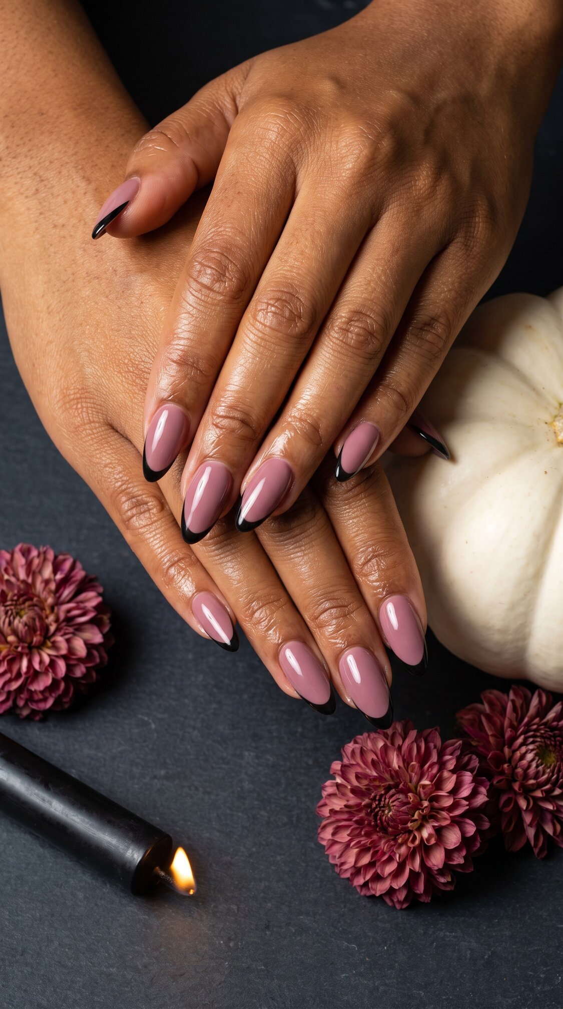 Hands with long, almond-shaped halloween nails acrylic, painted mauve with black tips, resting on a dark surface next to a white pumpkin, pink flowers, and a lit black candle.