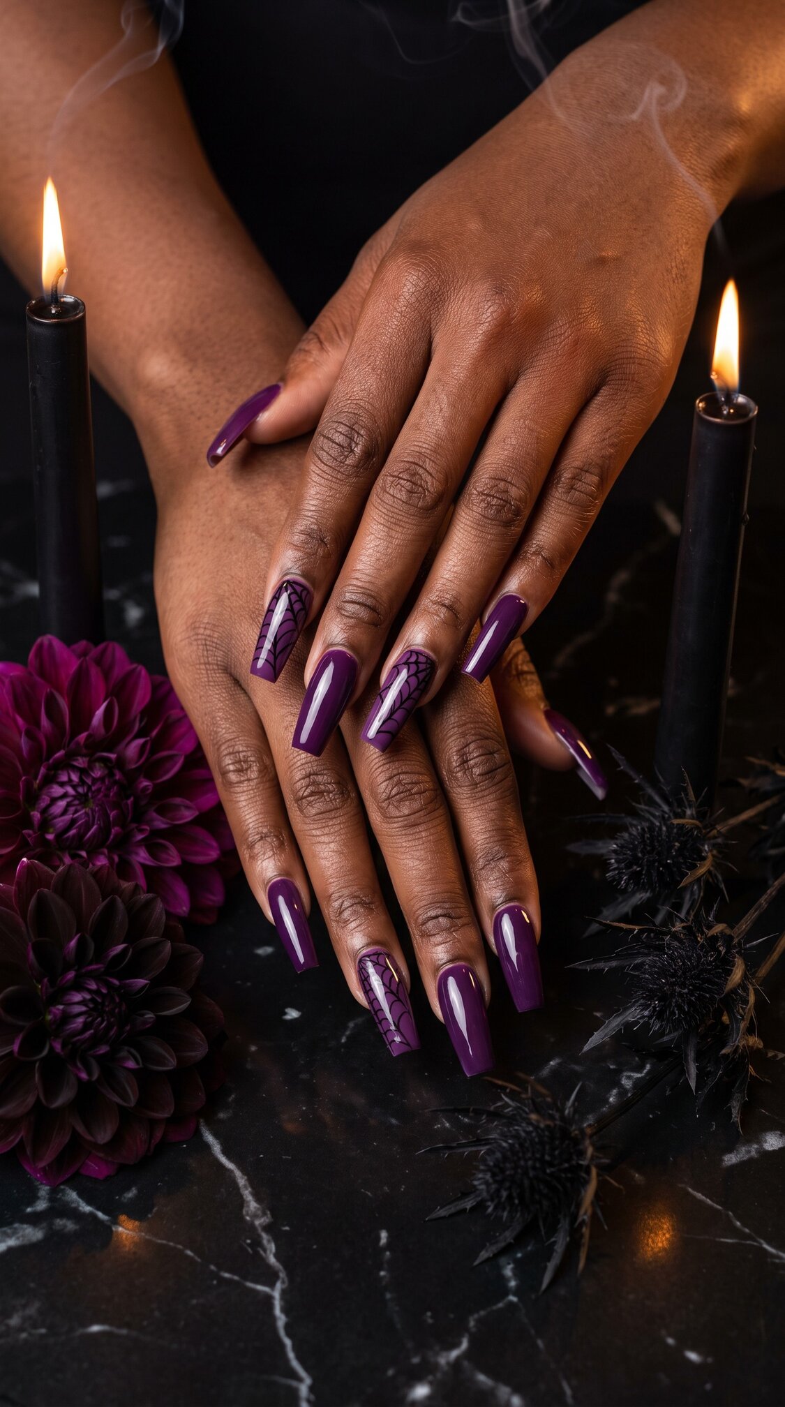 Two hands with long, glossy dark purple halloween nails acrylic rest on a black marble surface, surrounded by black candles, purple flowers, and black thistles. Smoke rises from the candles, creating a moody, elegant scene.