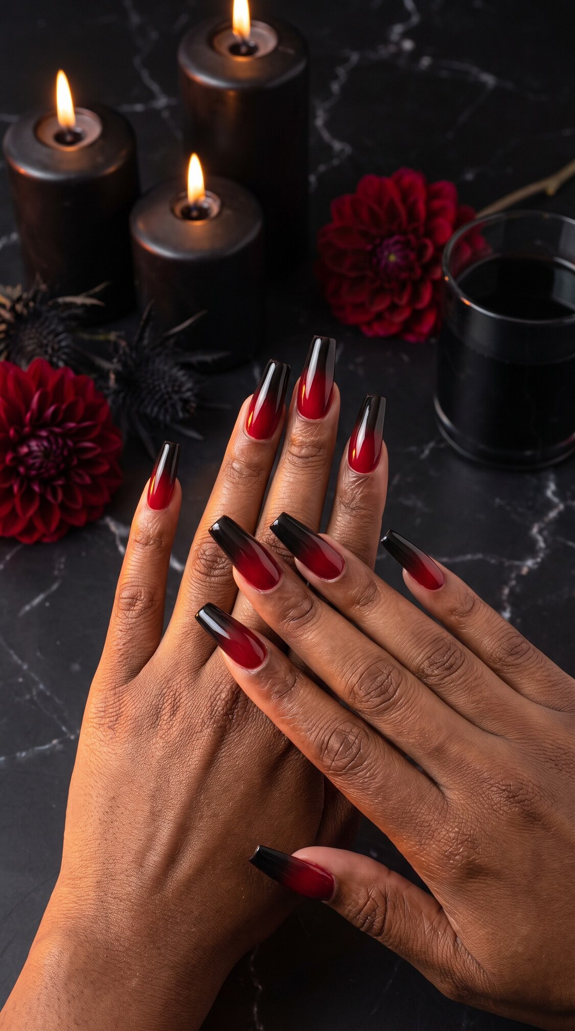 A pair of hands with long, red and black ombre halloween nails acrylic rests on a dark marble surface, surrounded by lit black candles, red flowers, and a glass of dark liquid.
