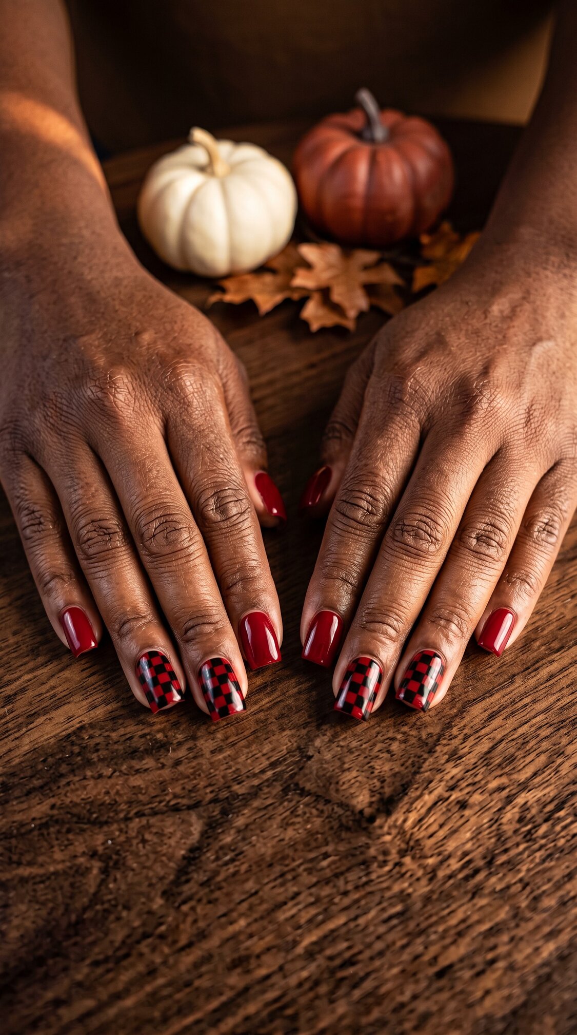 Two hands with red and plaid-patterned halloween nails acrylic rest on a wooden table. Small white and orange pumpkins and autumn leaves decorate the background.